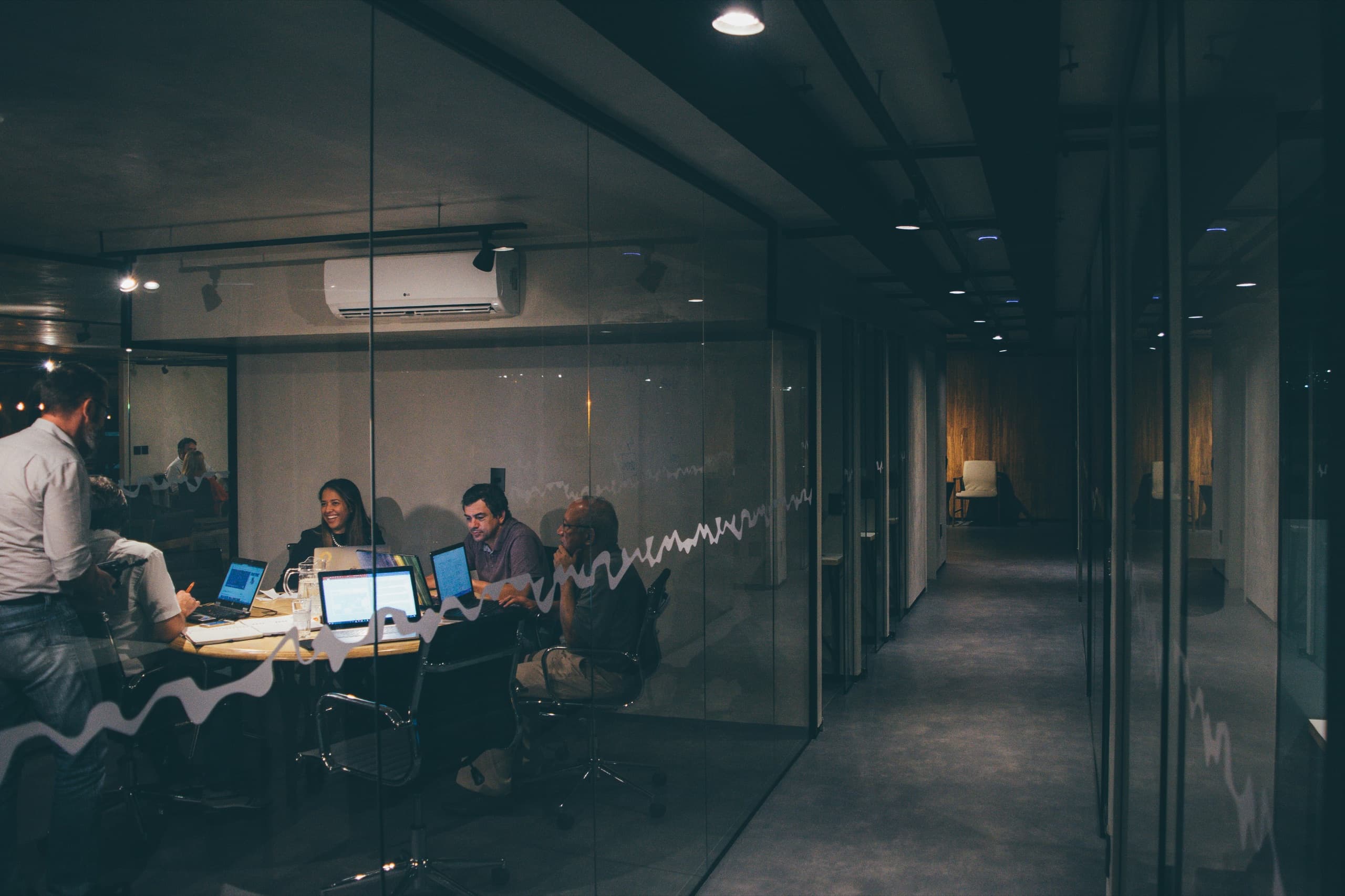 A team working on laptops inside a glass-walled meeting room at night